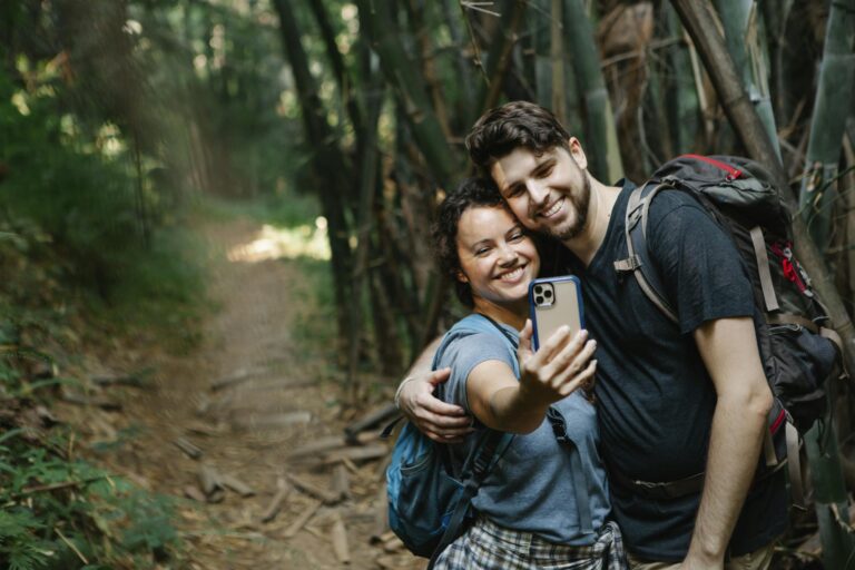 Cheerful young couple in casual clothes and backpacks cuddling and smiling while taking selfie on mobile phone during trekking in green forest