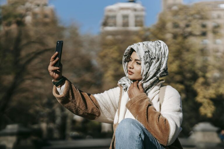 Gorgeous young Muslim female millennial in trendy outfit and hijab resting in autumn park and taking selfie on smartphone